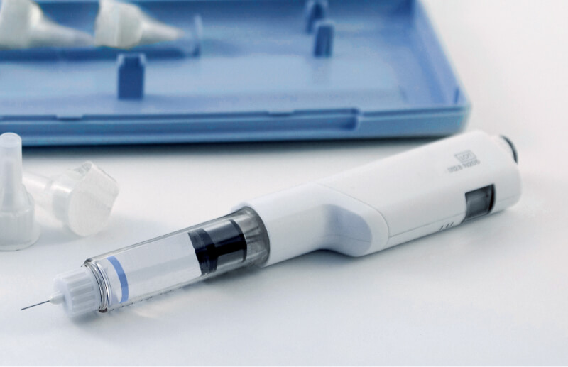 White insulin pen with an exposed needle and transparent cartridge lying on a white table with a blue tray in the background..
