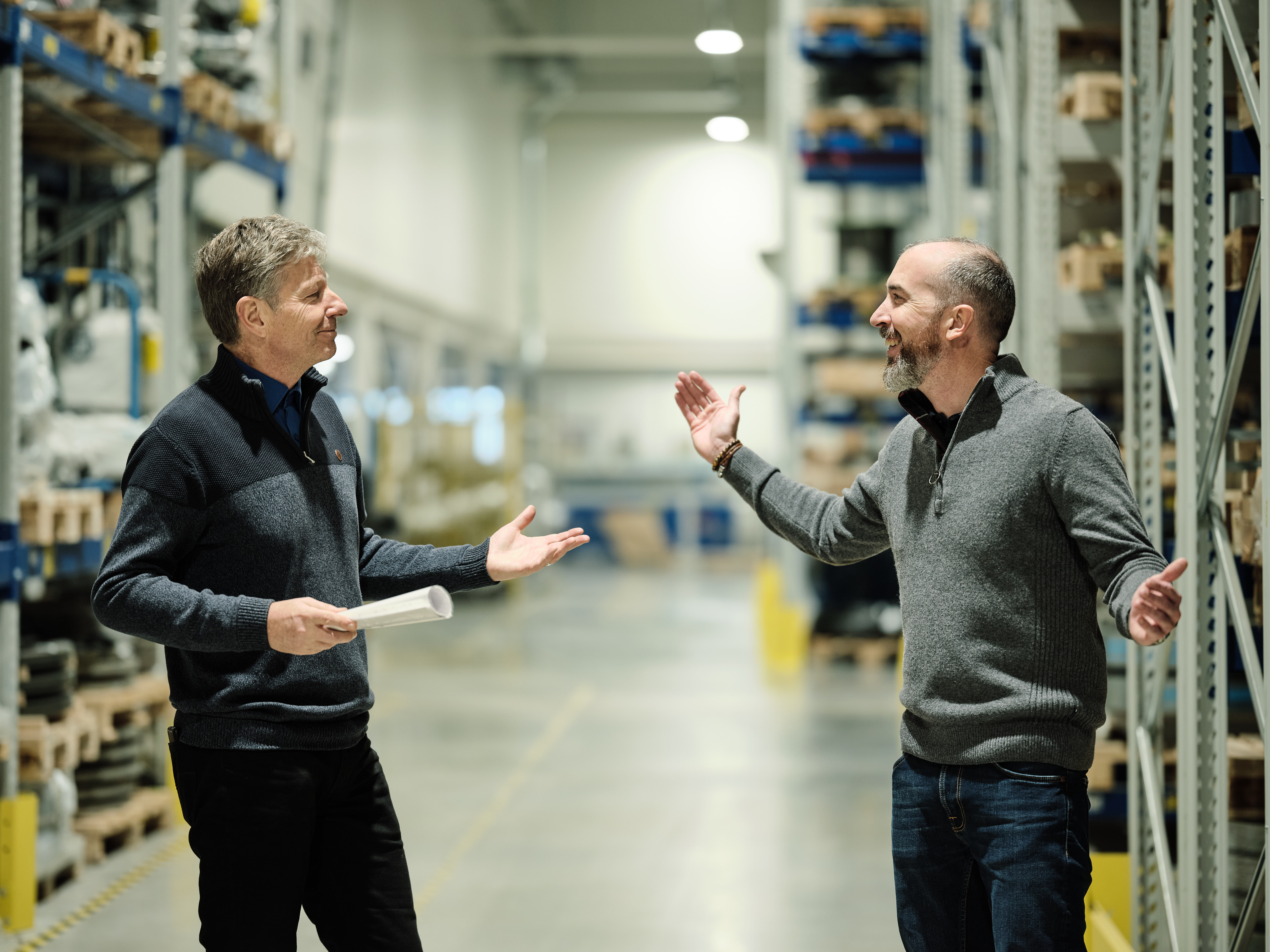 Zwei Männer in einem Lager, die lächeln und gestikulieren, während sie miteinander sprechen. Regale voller Kisten säumen die Wände unter heller industrieller Beleuchtung.