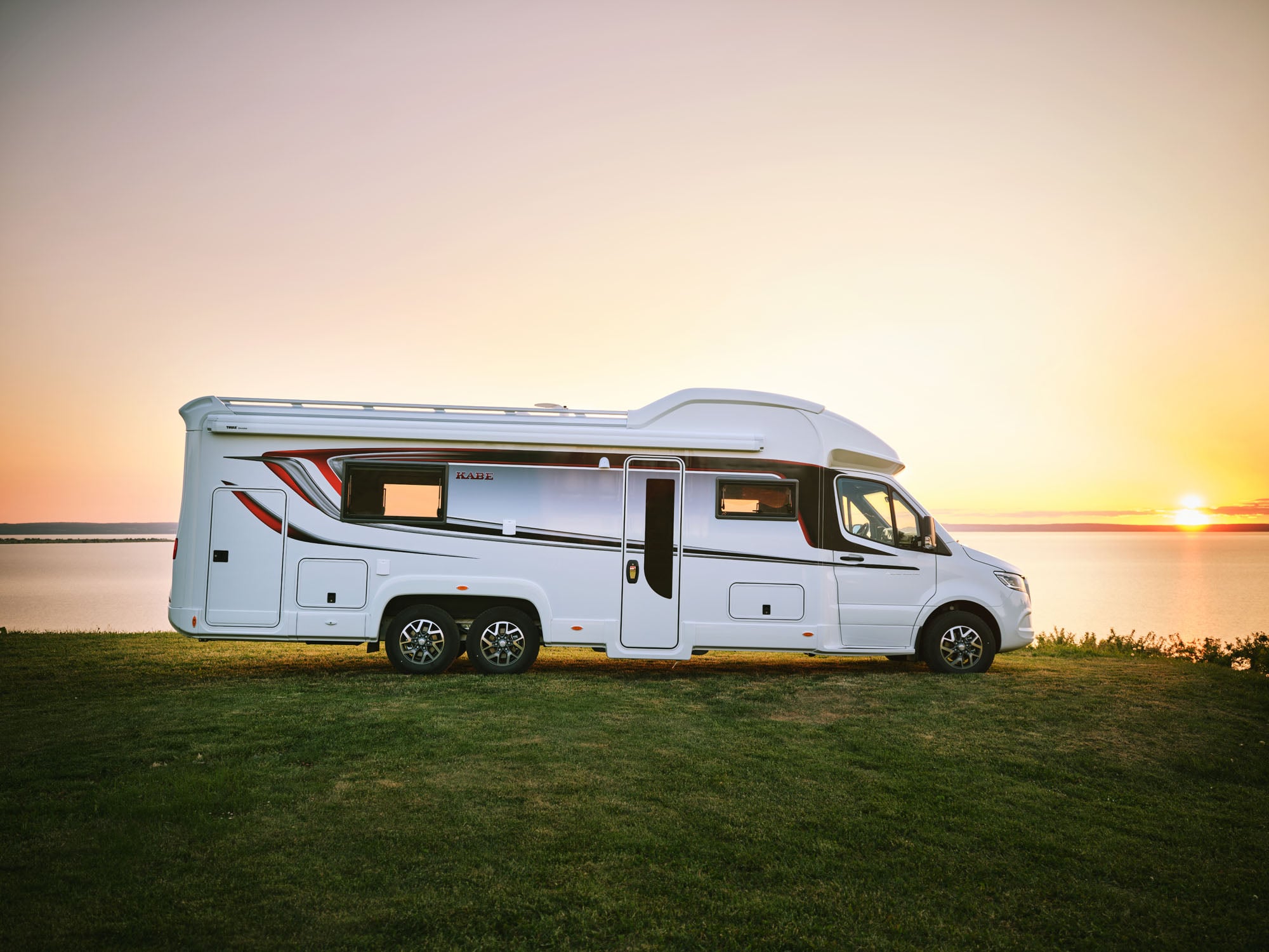 En campervan er parkeret ved en rolig sø ved solnedgang, med en blød orange og pink himmel.