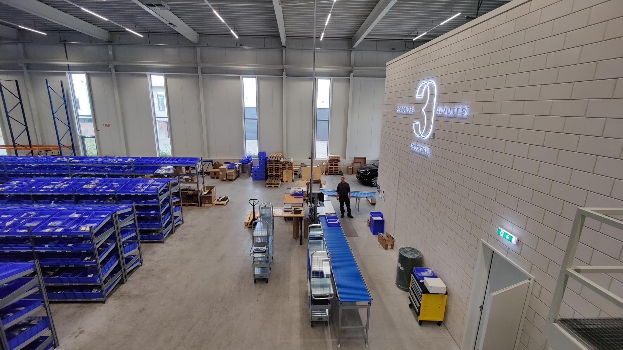 Eine geräumige Industriehalle mit Reihen blauer Kisten auf der linken Seite, Arbeitern, die in der Mitte Waren auf Förderbändern bearbeiten, und einem großen Logo an der rechten Wand.