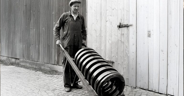 Schwarzweißfoto eines Mitarbeiters, der eine große Drahtfeder auf einem Handwagen bewegt.