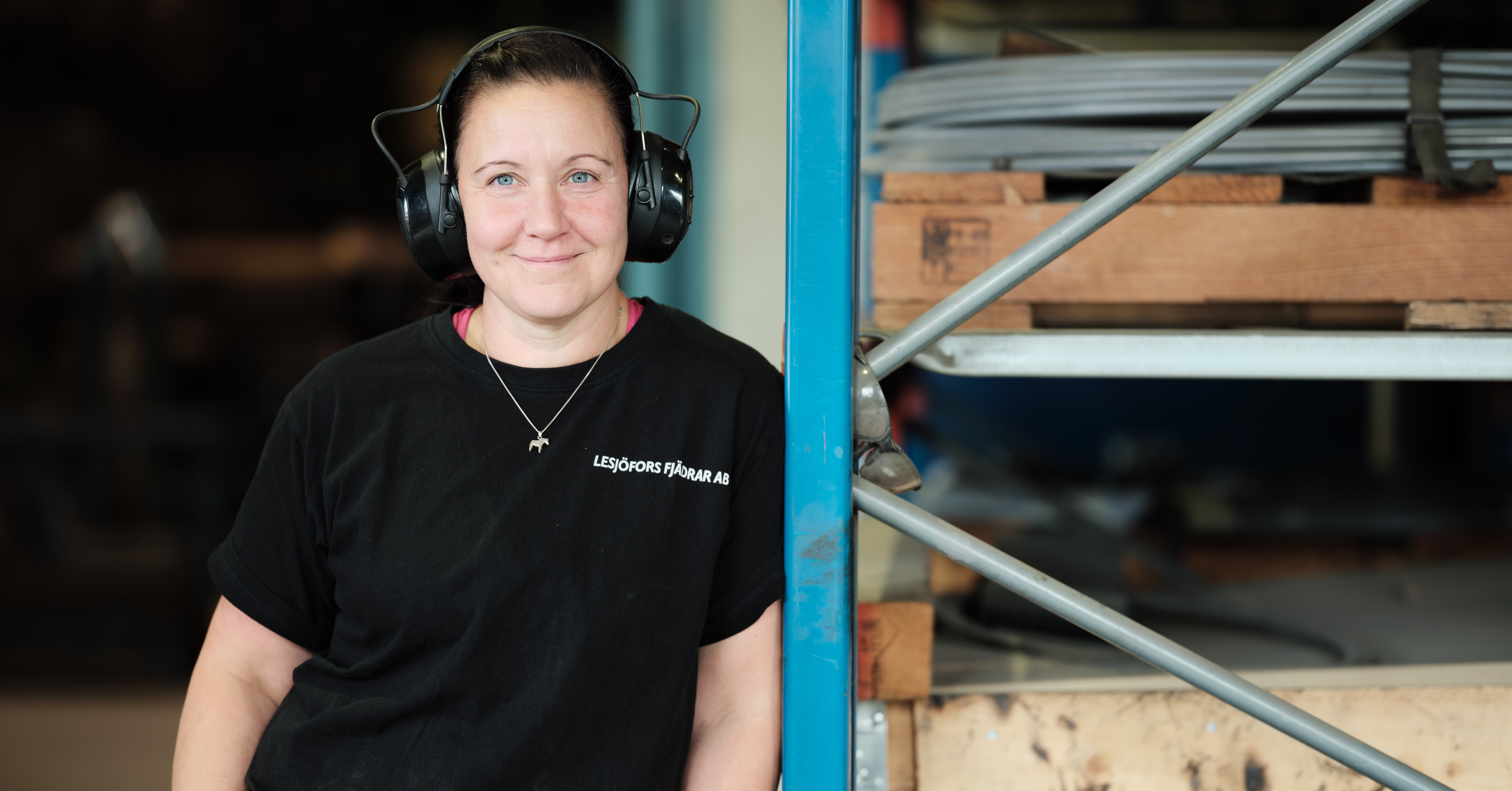 En person med kort hår, iført hovedtelefoner og en sort t-shirt, står smilende ved siden af industrielle hylder i et værkstedsmiljø.