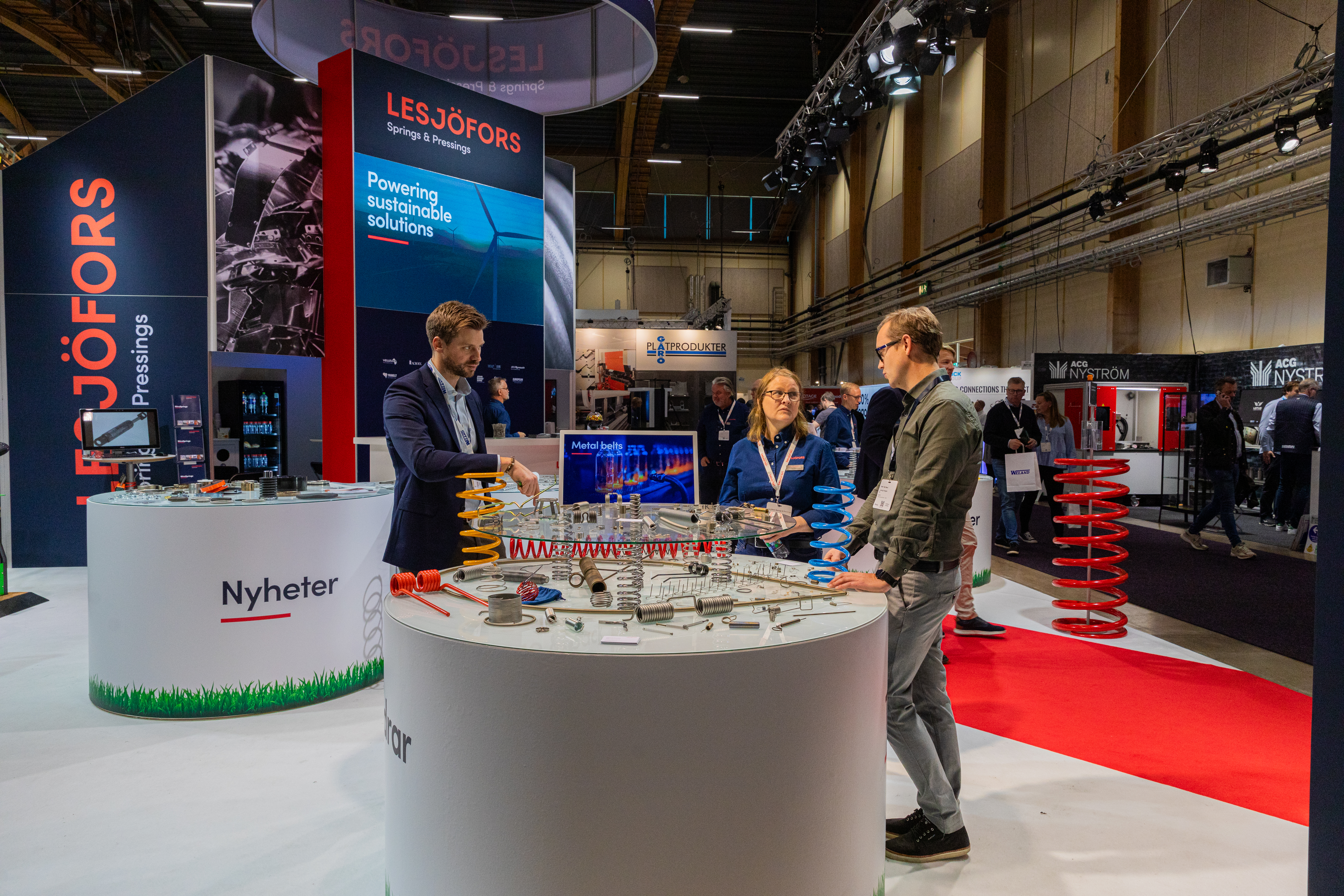 Drei Personen besprechen Produkte an einem Lesjöfors-Messestand, auf einem runden Tisch sind verschiedene Industriefedern und Metallkomponenten ausgestellt.