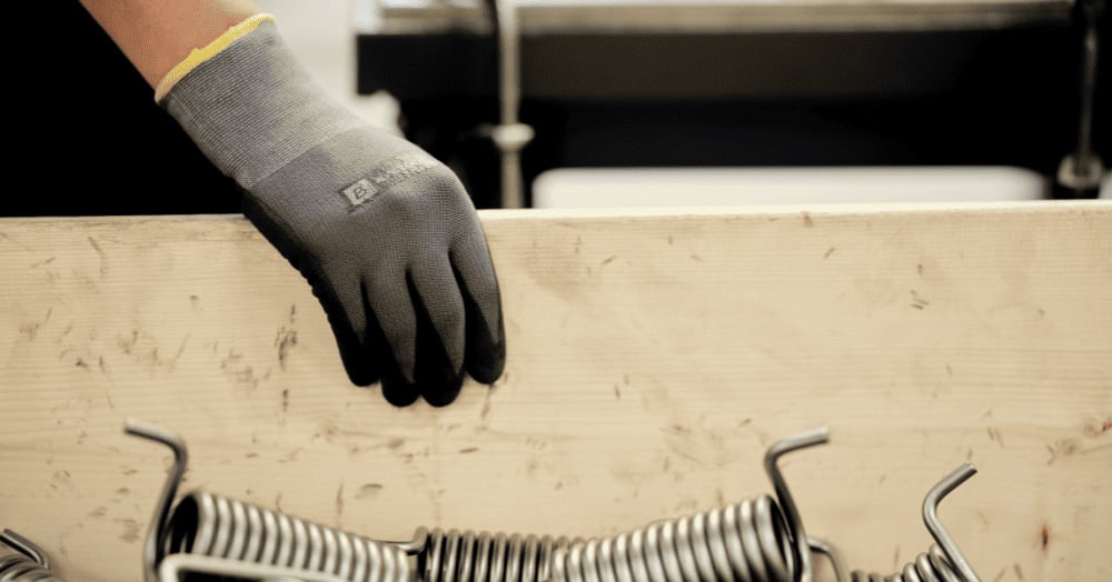 Eine behandschuhte Hand liegt auf einem Holzbrett, im Vordergrund befindet sich ein Haufen Metallfedern.
