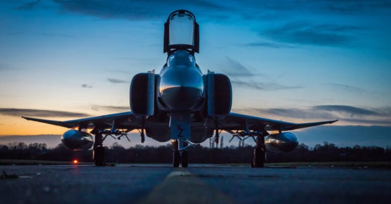 Ein Kampfjet steht in der Dämmerung auf einer Landebahn, als Silhouette vor einem dramatischen blau-orangen Himmel.