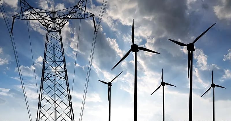 Ein großer Strommast und mehrere Windkraftanlagen stehen unter einem bewölkten Himmel und veranschaulichen Erzeugung und Verteilung elektrischer Energie.