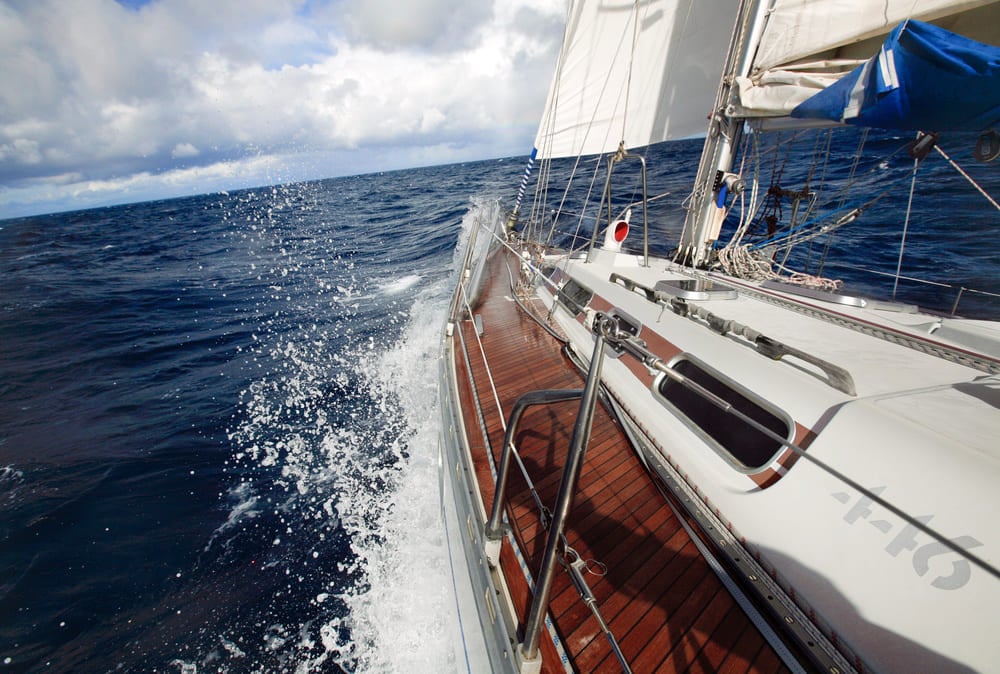 Onboard view of a sailing yacht moving through ocean waves, with water splashing along the side under a partly cloudy sky.