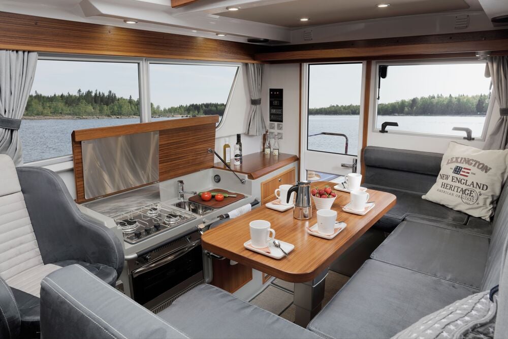 Boat cabin interior with dining area, kitchen, and large windows overlooking the water.