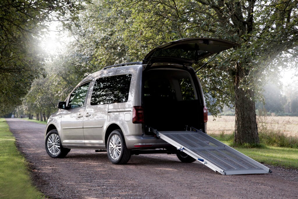 A silver minivan with the rear hatch open and a ramp extended, parked on a tree-lined road.