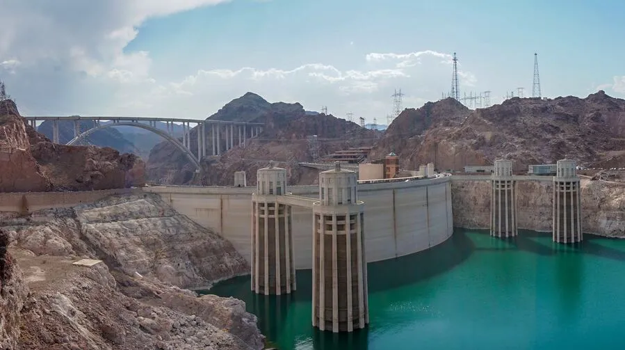 Große Betontalsperre mit Wasserreservoir, umgeben von felsiger Landschaft und einer Brücke im Hintergrund.