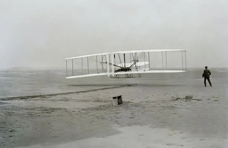 Early Wright brothers-style biplane flying low over a sandy beach with a person nearby.