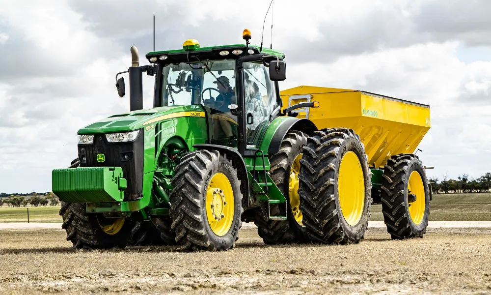 Ein großer grüner Traktor zieht einen gelben Anhänger über Ackerland unter einem bewölkten Himmel.