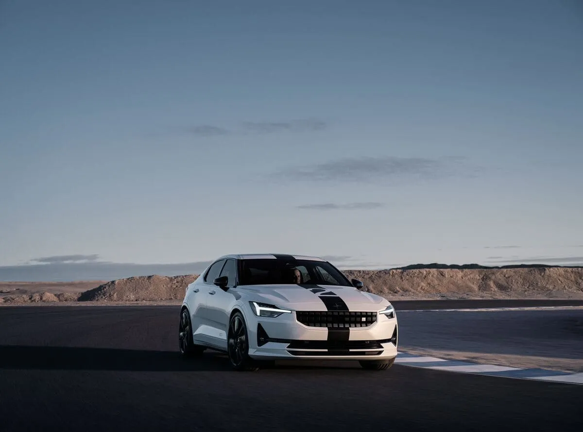 A white Polestar 2 BST Edition 270 electric performance car with a black racing stripe, photographed from the front on a racing circuit against a desert landscape and blue sky at dusk.