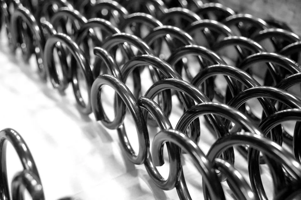 Nahaufnahme von schwarzen Metallspiralen in einem spiralförmigen Muster, das ein sich wiederholendes, geometrisches Design erzeugt. Die glänzende Oberfläche bildet einen Kontrast zu einem verschwommenen, hellen Hintergrund.