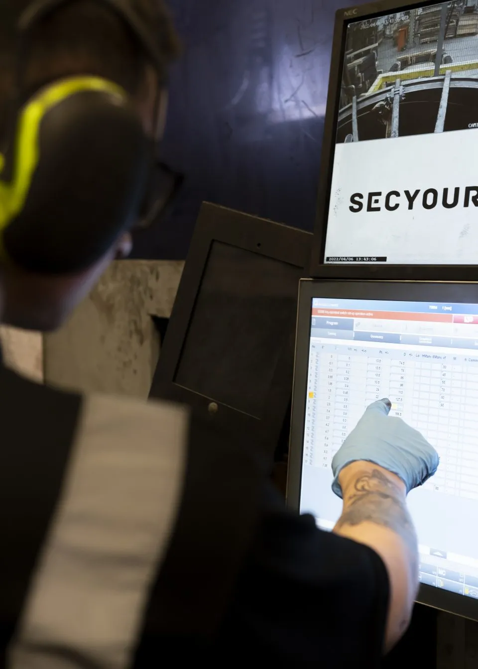 Unscharfes Bild einer Person, die eine Brille und Gehörschutz trägt und einem Laptop zugewandt ist. Der Hintergrund ist undeutlich und vermittelt Unsicherheit oder Konzentration.