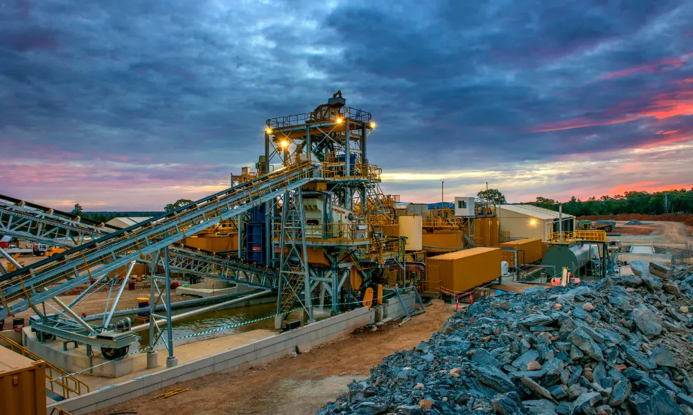 Beleuchtete Mineralaufbereitungsanlage mit Förderbandsystemen und aufgeschüttetem Gestein in der Dämmerung, vor einem dramatischen Sonnenuntergangshimmel.