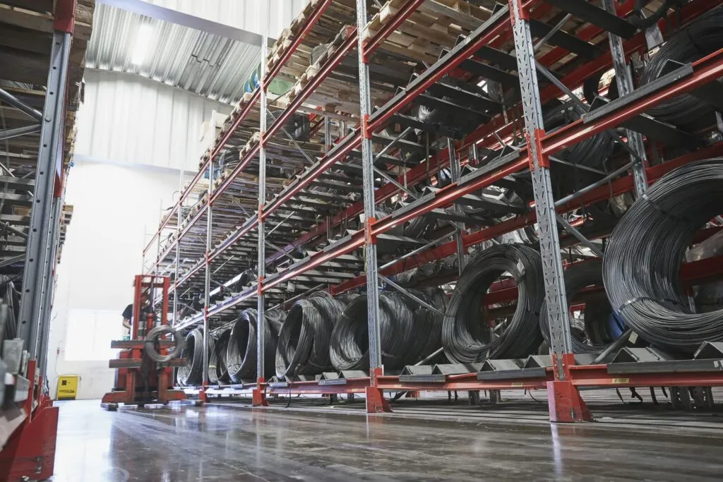 Innenansicht eines großen Industrielagers mit hohen roten Metallregalsystemen, in denen auf mehreren Ebenen zahlreiche Coils aus Walzdraht gelagert sind, mit einem polierten Betonboden im Vordergrund.