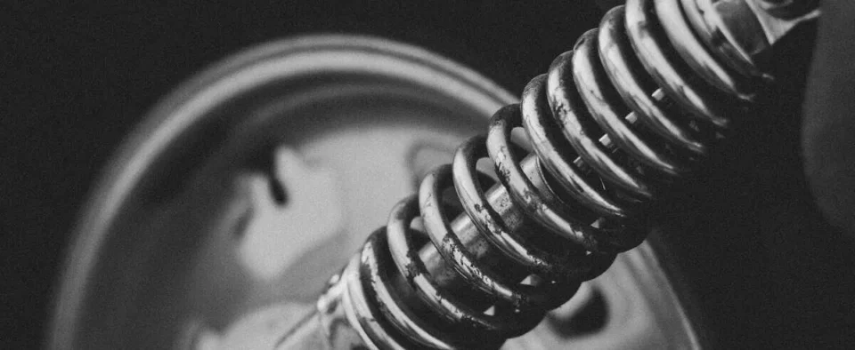 Black-and-white close-up of a motorcycle suspension coil spring with wheel in background.