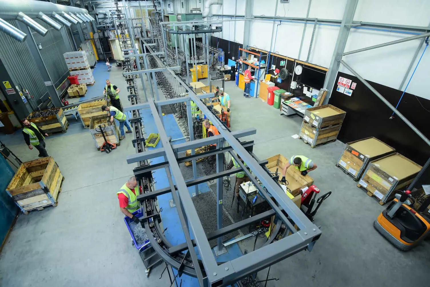 An overhead view of a busy powder coating factory floor in Redruth, Cornwall, with workers in hi-vis vests operating conveyor systems and handling wooden crates of springs.
