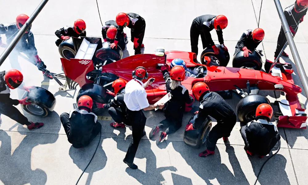 Die Boxencrew ist in voller Aktion und wechselt während eines Boxenstopps die Reifen an einem roten Rennwagen. Die Crewmitglieder in roten Helmen arbeiten effizient und schaffen eine dynamische, intensive Szene.