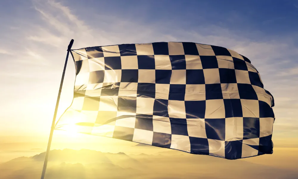 Eine karierte Zielflagge weht im Vordergrund und wird von einem warmen Sonnenuntergang beleuchtet. Der Himmel ist golden und vermittelt eine siegreiche und triumphale Stimmung.