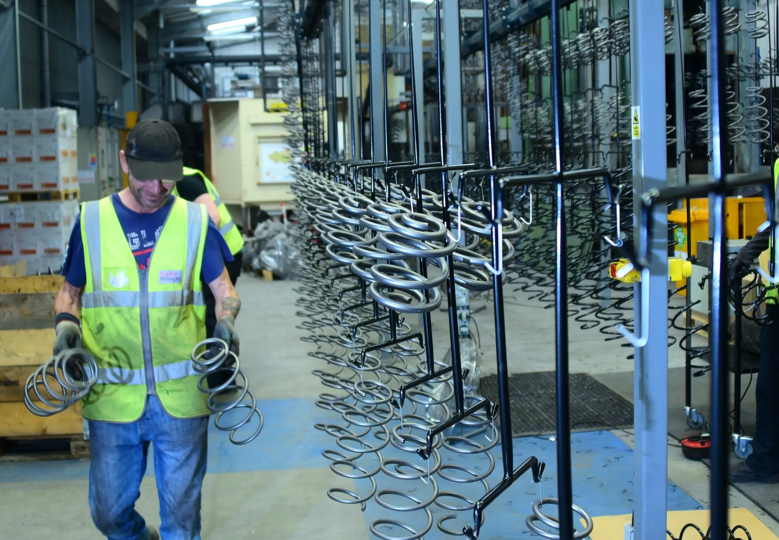 Racks of stainless steel springs sit in a factory while a man walks past holding two heavy-duty springs.
