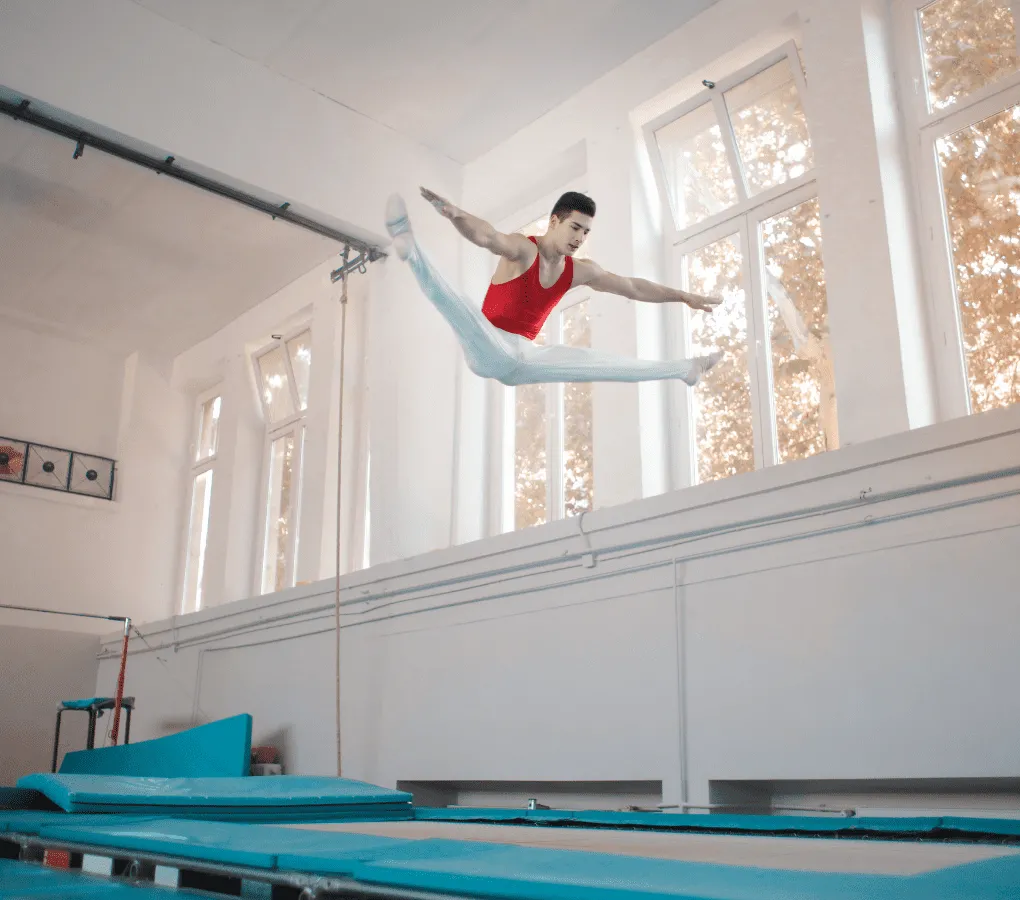 Ein männlicher Trampolinturner in rotem Trikot und weißer Hose zeigt einen Pike-Sprung hoch über einem blauen Trampolin in einer Turnhalle.