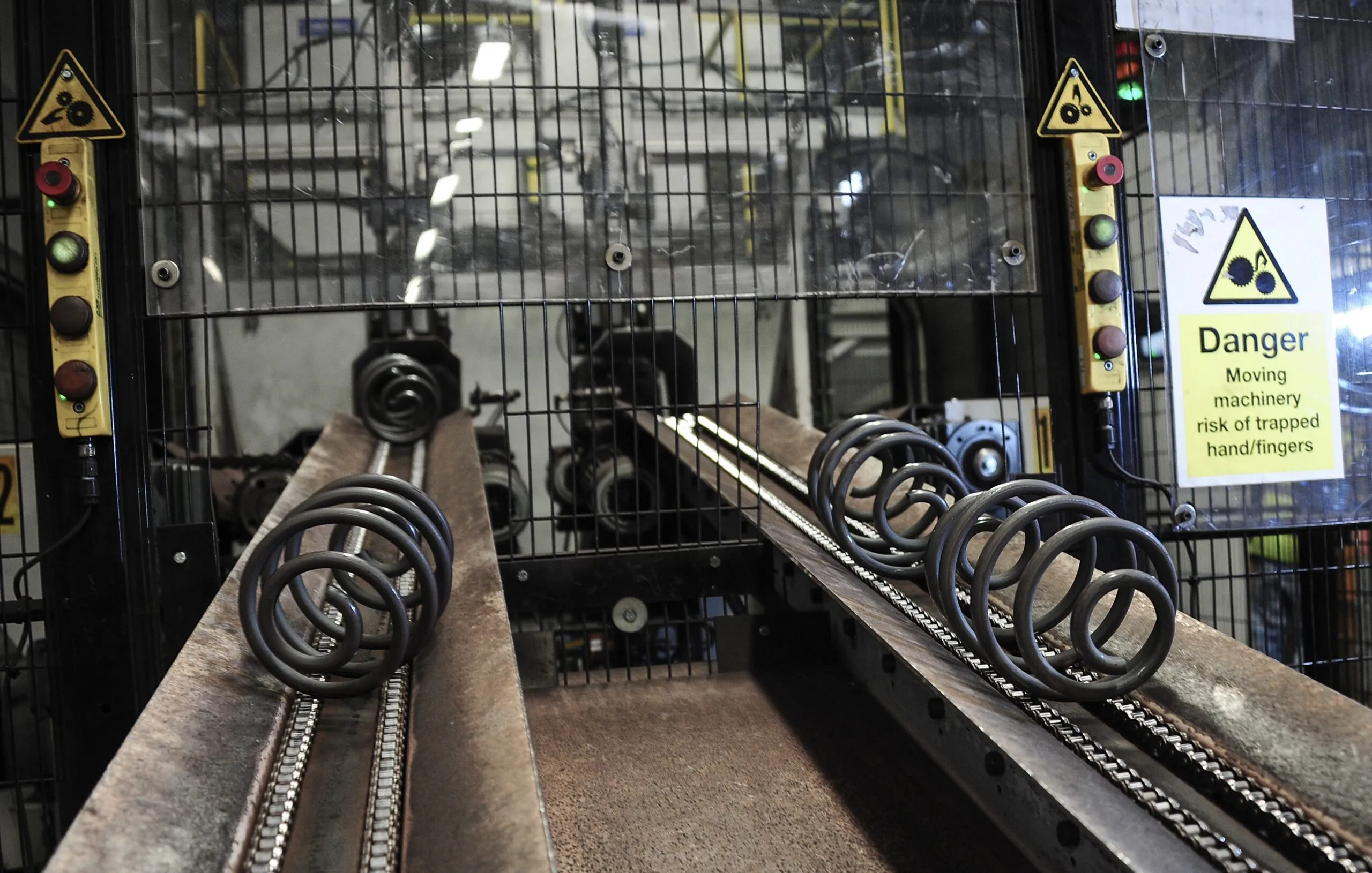 Dark metal coil springs transported along parallel conveyor tracks inside a guarded machine.