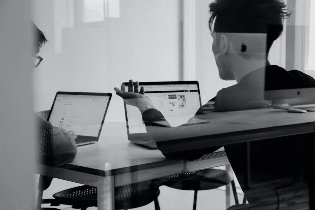 Zwei Personen arbeiten am Schreibtisch mit Laptops an der Auslegung von Drahtfedern. Eine Person gestikuliert während der Diskussion in Richtung der Bildschirme, fotografiert in Schwarzweiß.