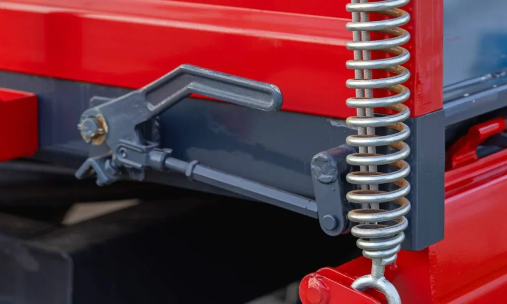 Nahaufnahme einer silberfarbenen Zugfeder mit Hakenende, vertikal an roter und grauer Schwermaschine montiert, neben einem metallischen Verriegelungsmechanismus.
