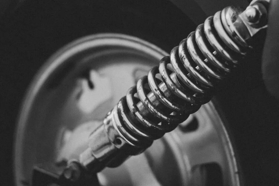 Black-and-white close-up of a motorcycle suspension spring with wheel in background.