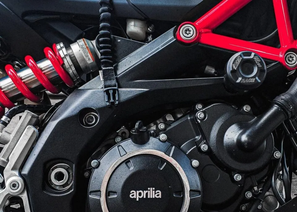 Close-up of Aprilia motorcycle rear suspension with red coil spring and engine components.