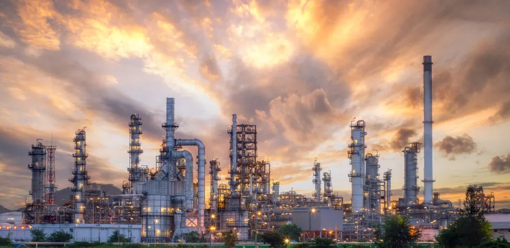 Ein beleuchteter Raffineriekomplex, bei Sonnenuntergang fotografiert, mit Türmen, Rohrleitungen und Industrieanlagen vor einem dramatischen orange-violetten Himmel.