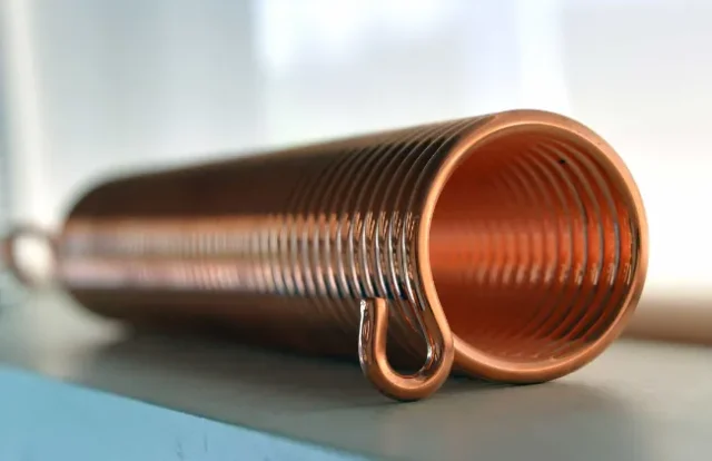 Nahaufnahme einer Kupferspirale mit spiralförmigen Rillen, die horizontal auf einer grauen Oberfläche liegt. Die Beleuchtung hebt ihre glänzende, metallische Struktur hervor.