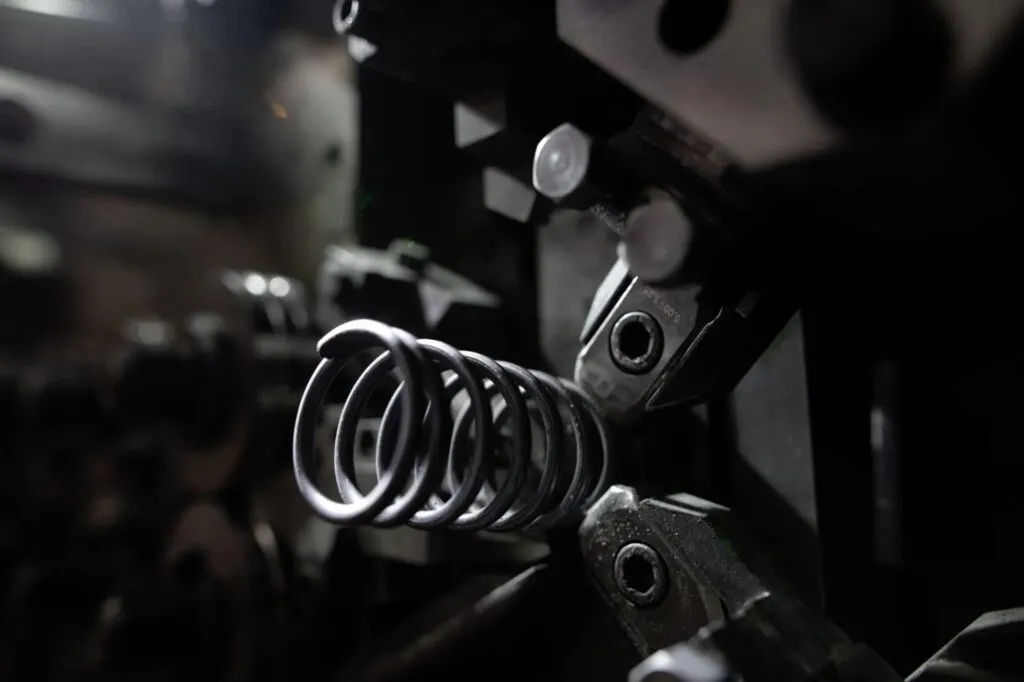 Nahaufnahme einer Stahl-Drahtfeder, die in einer industriellen Federwindemaschine geformt wird, mit sichtbaren mechanischen Komponenten und Werkzeugen im dunklen, unscharfen Hintergrund.