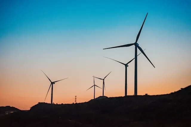 Silhouetted wind turbines on a hillside at sunset with an orange and blue sky.