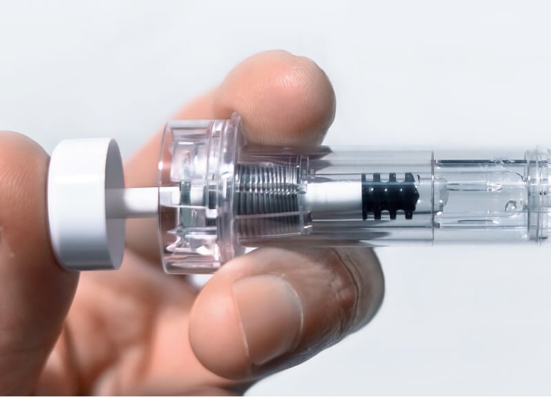 Eine Hand hält ein transparentes spritzenähnliches Gerät mit einem sichtbaren Federmechanismus im Inneren. Das Bild vermittelt einen klinischen, präzisen und sauberen Eindruck.