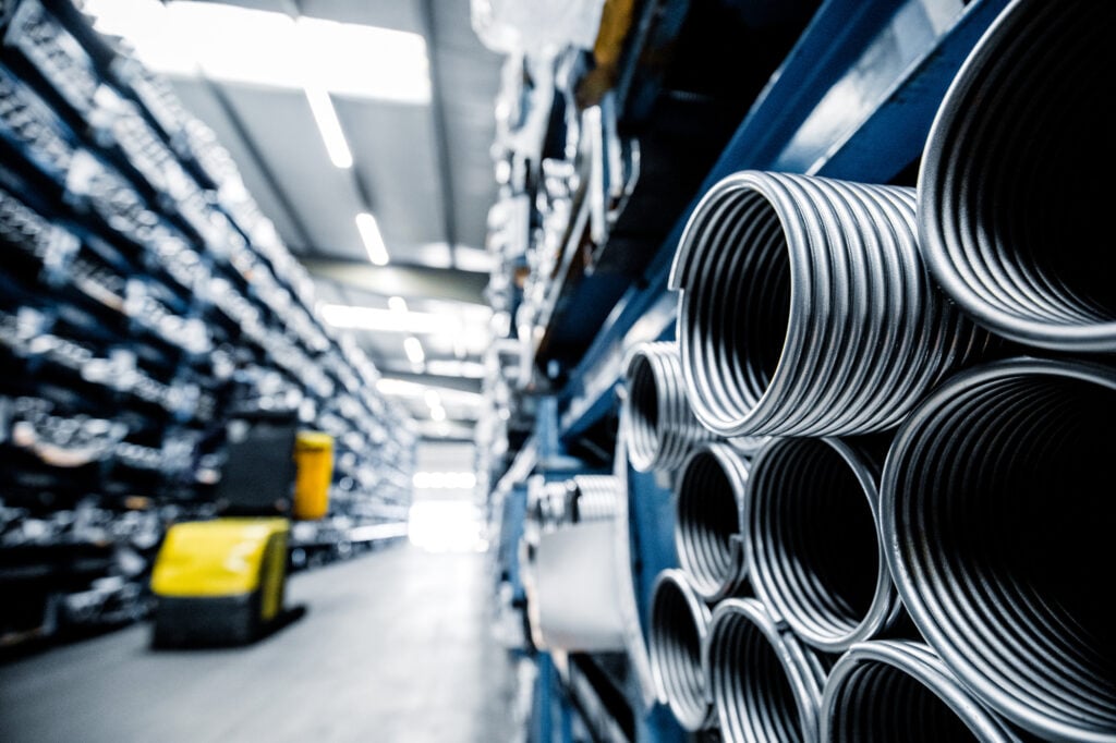 Ein hell beleuchteter Gang in einem Industrie-Lager mit gestapelten Metallrohren auf der rechten Seite und einem gelben Gabelstapler in der Ferne.
