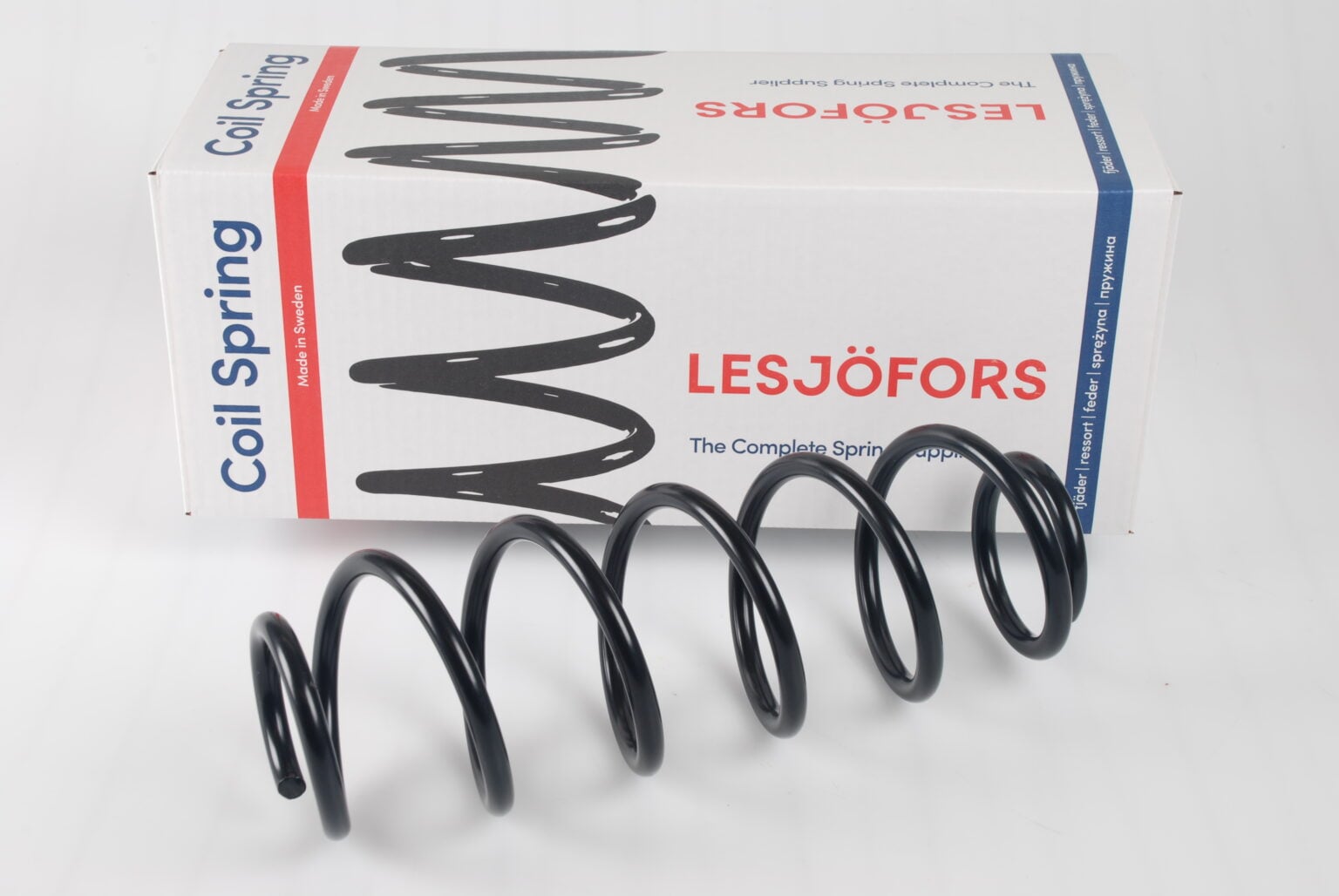 Nahaufnahme einer schwarzen Schraubenfeder vor einer weißen Box mit der Aufschrift „Lesjöfors“ und roten sowie blauen Akzenten. Die Feder ist geneigt und wirft einen Schatten.