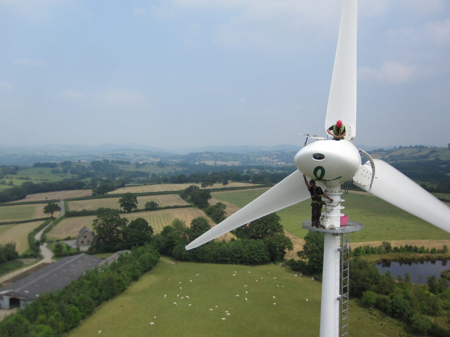 Eine Person steht auf einer großen weißen Windturbine vor einer Kulisse aus üppig grünen Feldern und entfernten Hügeln unter einem klaren blauen Himmel.
