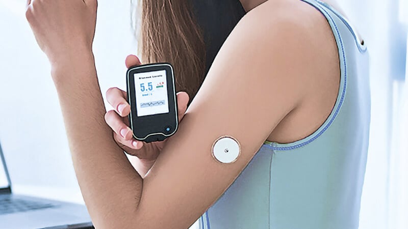 En kvinde holder en blodsukkermåler, der viser "5,5", ved siden af en sensor på armen, hvilket antyder diabetesbehandling. Hun bærer en lyseblå top.