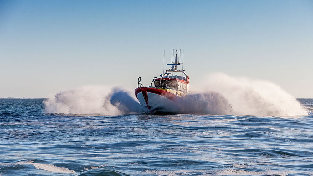 Ein rot-weißes Rettungsboot rast über den Ozean und erzeugt große Wellen und Gischt vor einem klarblauen Himmel - das vermittelt Dringlichkeit und Action.