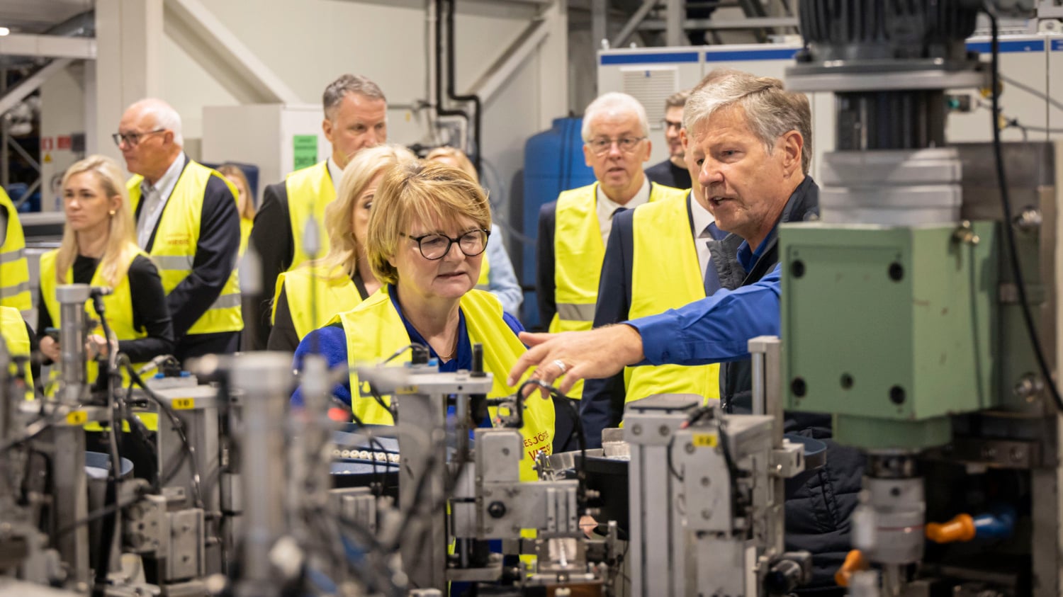 Eine Gruppe von Menschen in einer Fabrikumgebung, die gelbe Sicherheitswesten tragen, beobachtet einen Mann, der Maschinen vorführt.
