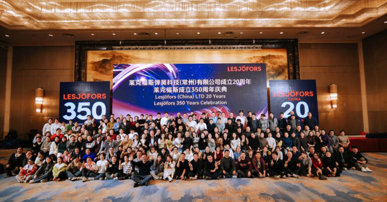 Eine große Gruppe von Menschen posiert drinnen für ein festliches Foto. Hinter ihnen hängen Banner mit der Aufschrift „Lesjöfors 350“ und „Lesjöfors (China) LTD 20 Years Celebration“.