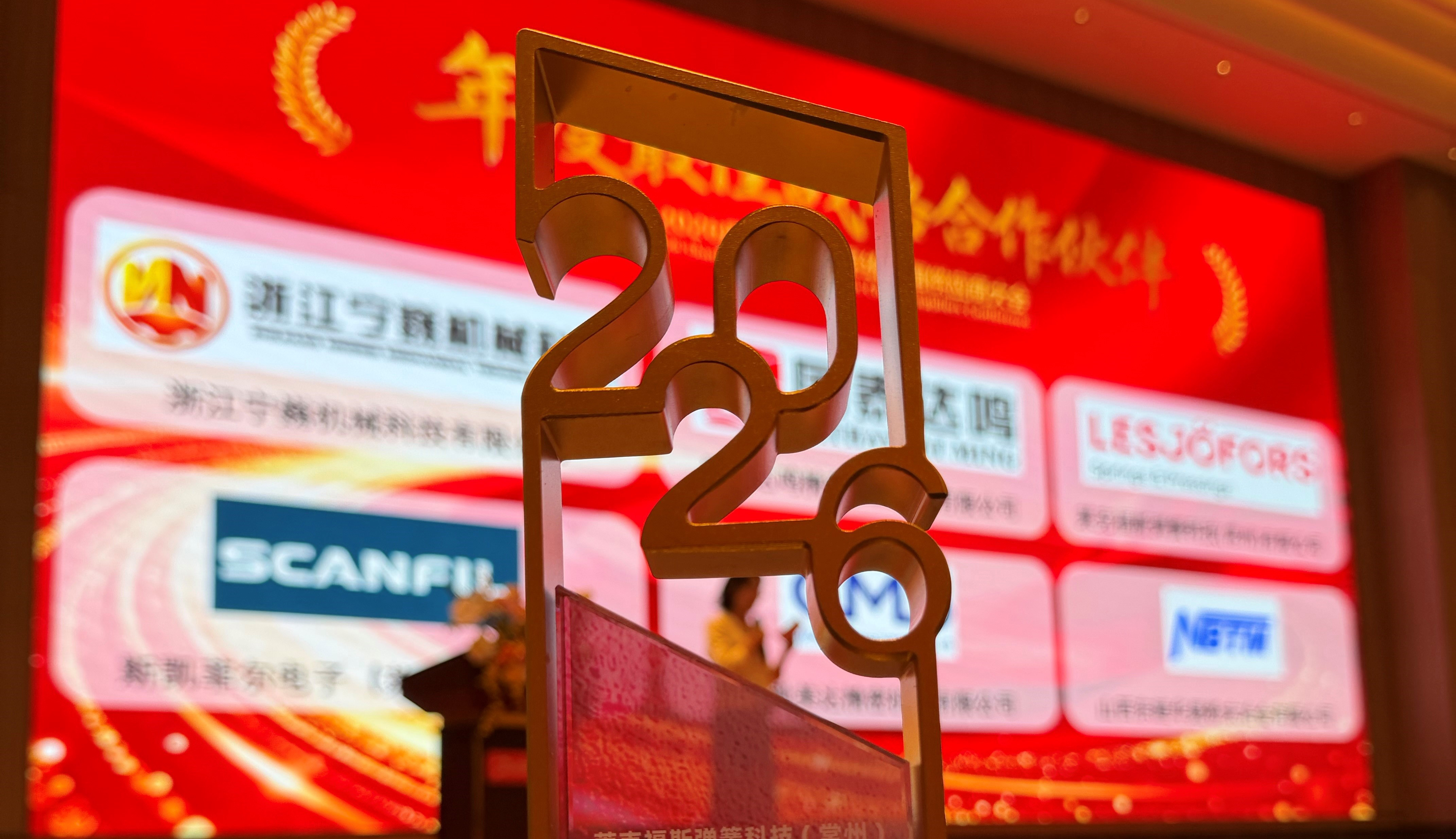 Nahaufnahme einer metallischen „2022“-Auszeichnung im Vordergrund, vor einem roten Veranstaltungsbanner mit chinesischem Text, mehreren Firmenlogos (darunter Lesjöfors und Scanfil) und einer unscharfen Person an einem Rednerpult im Hintergrund.