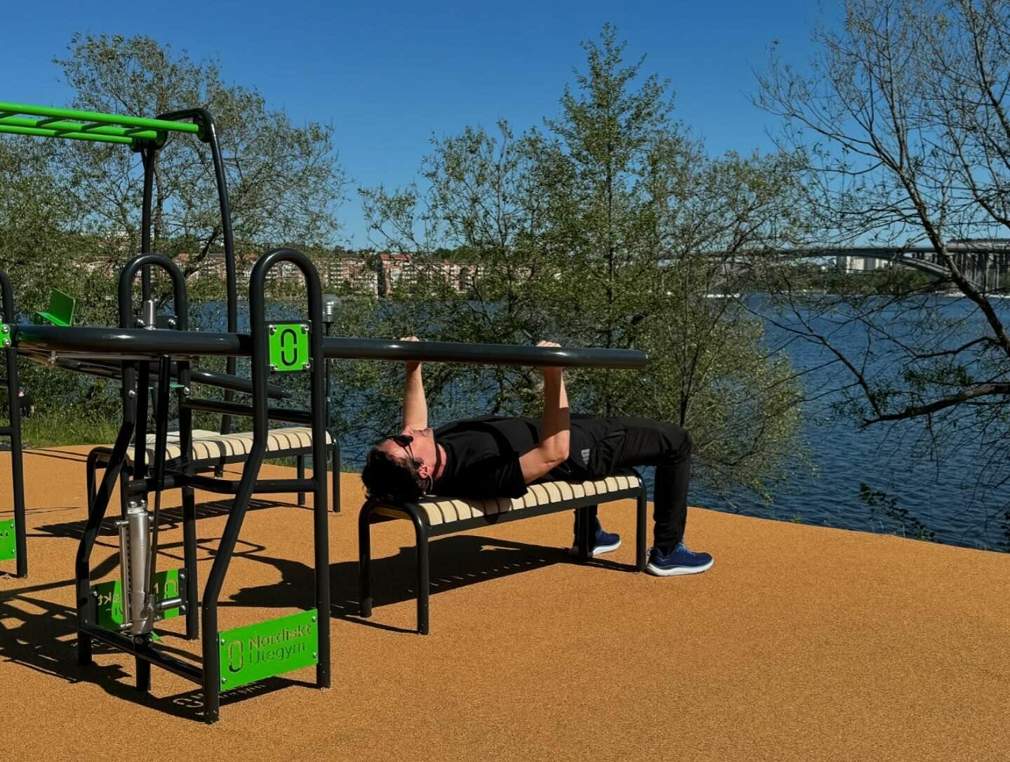 Eine Person trainiert an Outdoor-Fitnessgeräten an einem Fluss, umgeben von Bäumen unter einem klaren blauen Himmel.