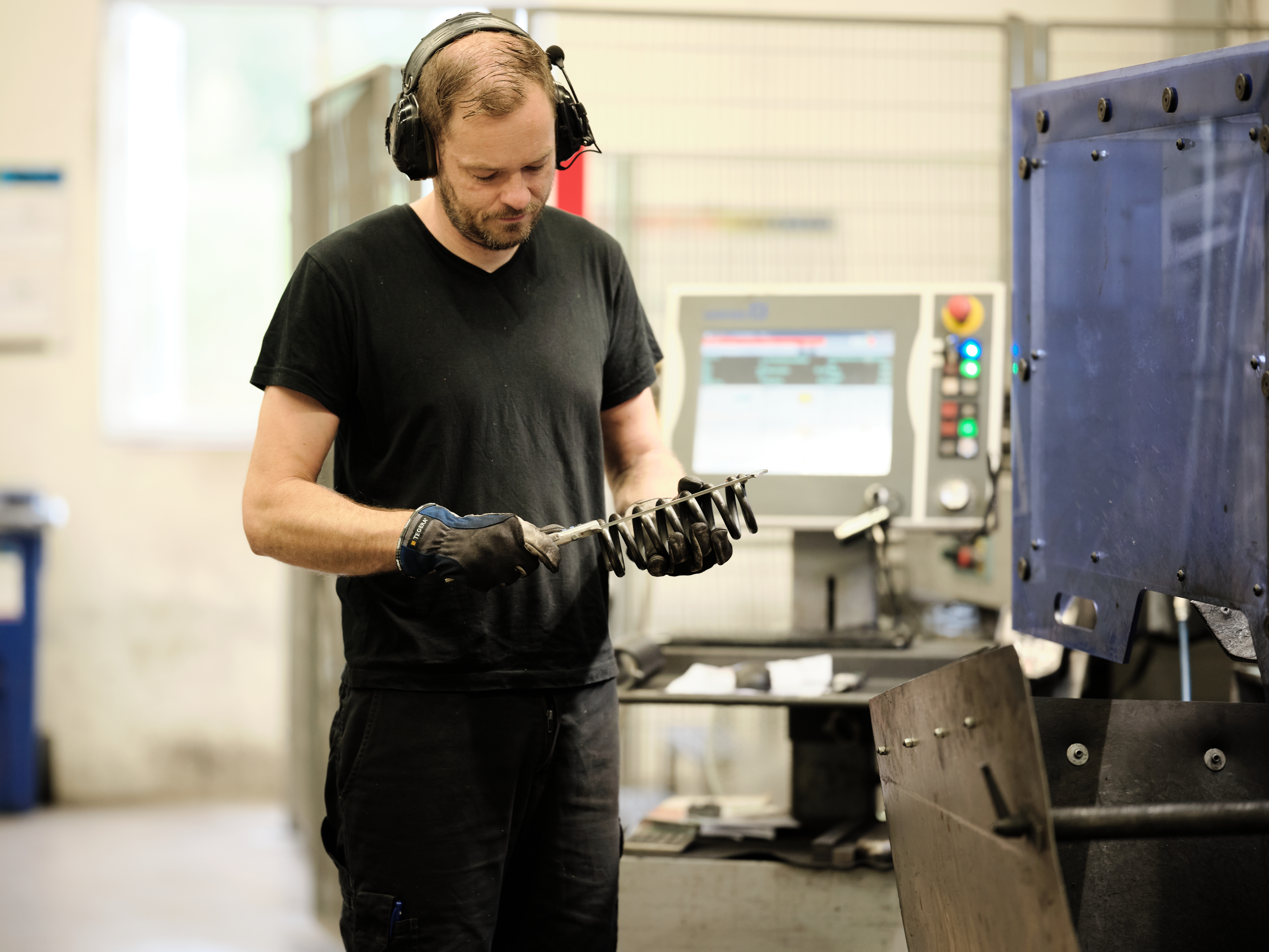 Lesjöforsin työntekijä, jolla on kuulosuojaimet ja käsineet, tarkastaa mustaa puristusjousta tehdasympäristössä.