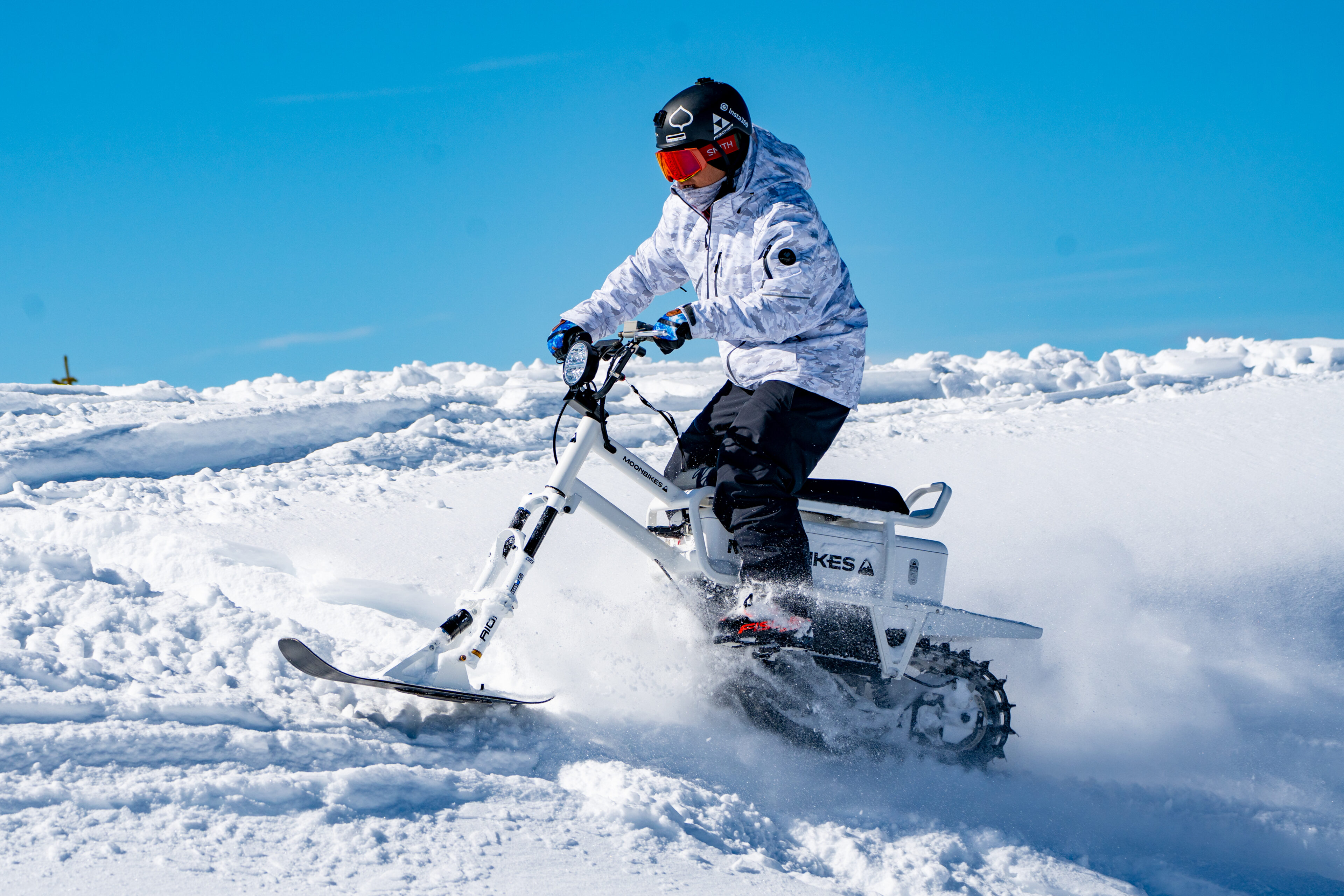 Eine Person fährt mit einem Schneefahrrad über eine frische Schneedecke unter klarem blauem Himmel, gekleidet in Winterausrüstung und mit einem roten Helm.