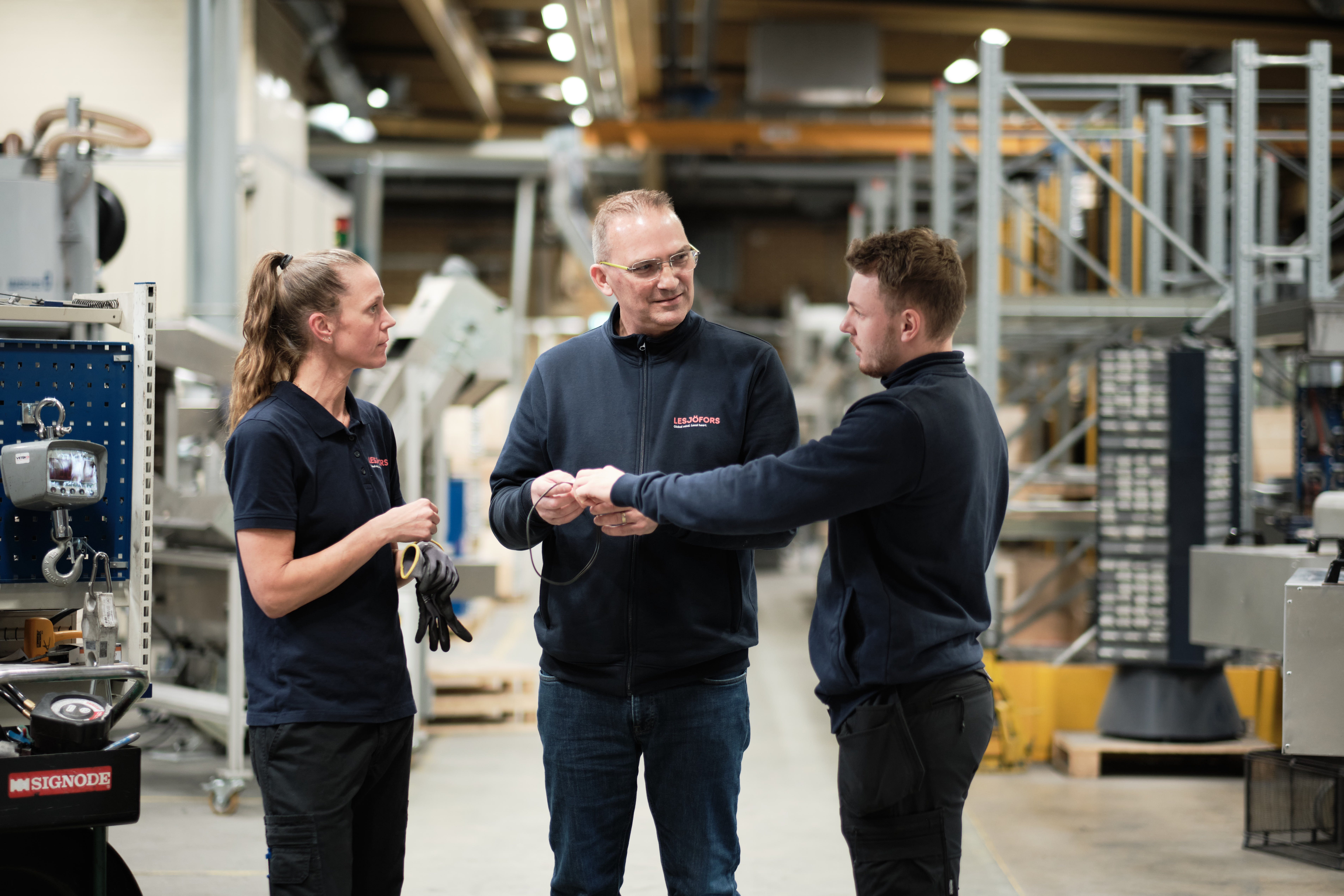 Drei Personen in einer Fabrikumgebung, zwei junge Personen hören aufmerksam einer älteren Person zu. Maschinen und Ausrüstung sind im Hintergrund sichtbar.