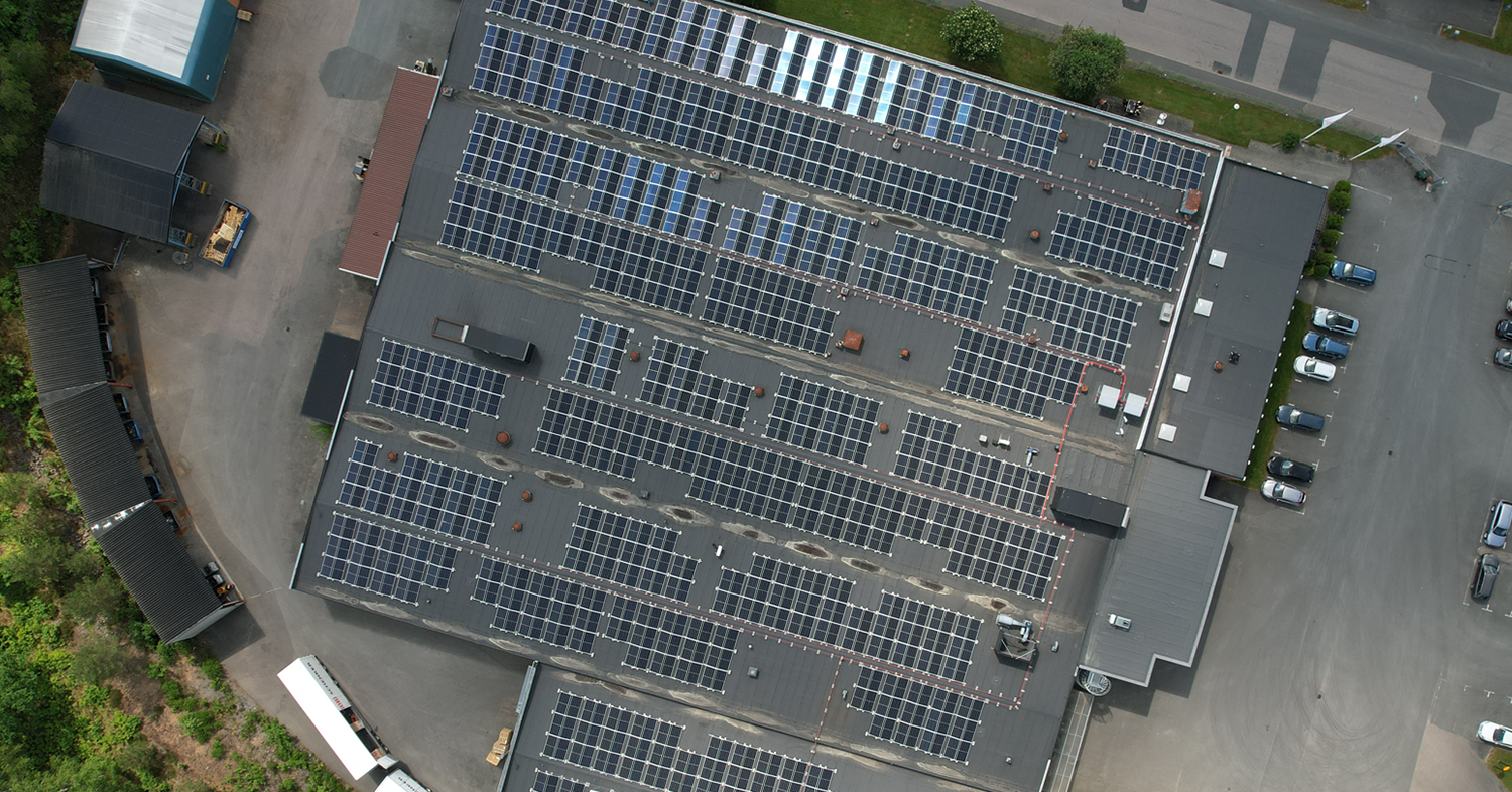 Luftaufnahme eines großen Industriedachs, das mit zahlreichen Solarmodulen bedeckt ist, umgeben von Grünflächen und Parkplätzen.